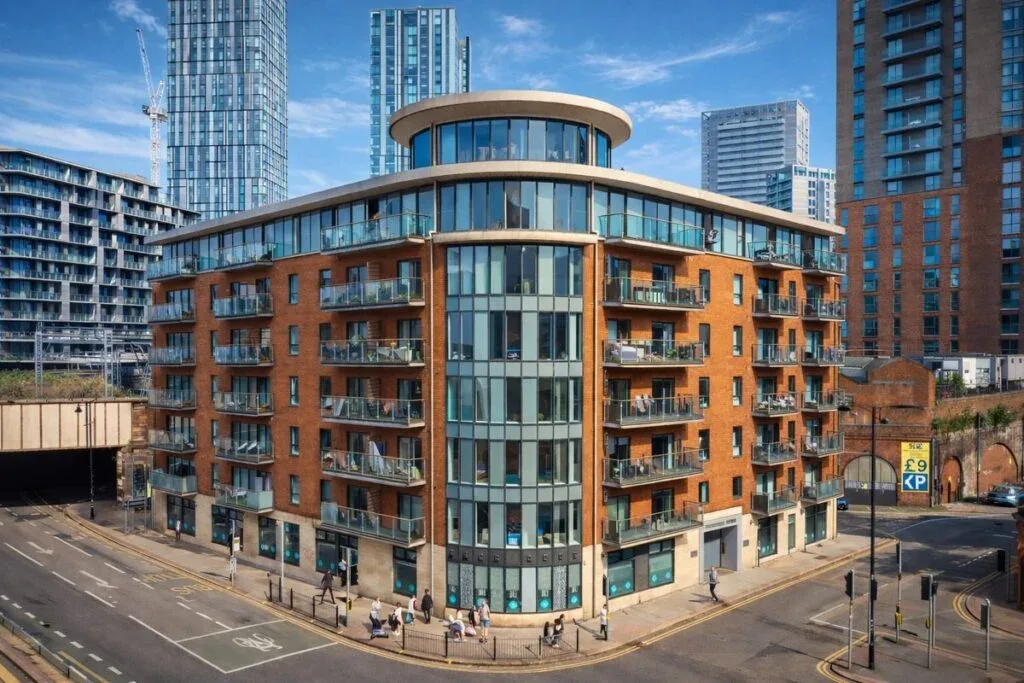 Photograph of the Jack Ross Chartered Accountants office building in central Manchester: a modern multi-storey red-brick and glass building with a curved corner section, glass balconies on each floor, glass shopfronts at street level, and a rounded cream-and-glass rooftop pavilion, photographed under blue sky with the central Manchester skyline visible behind.